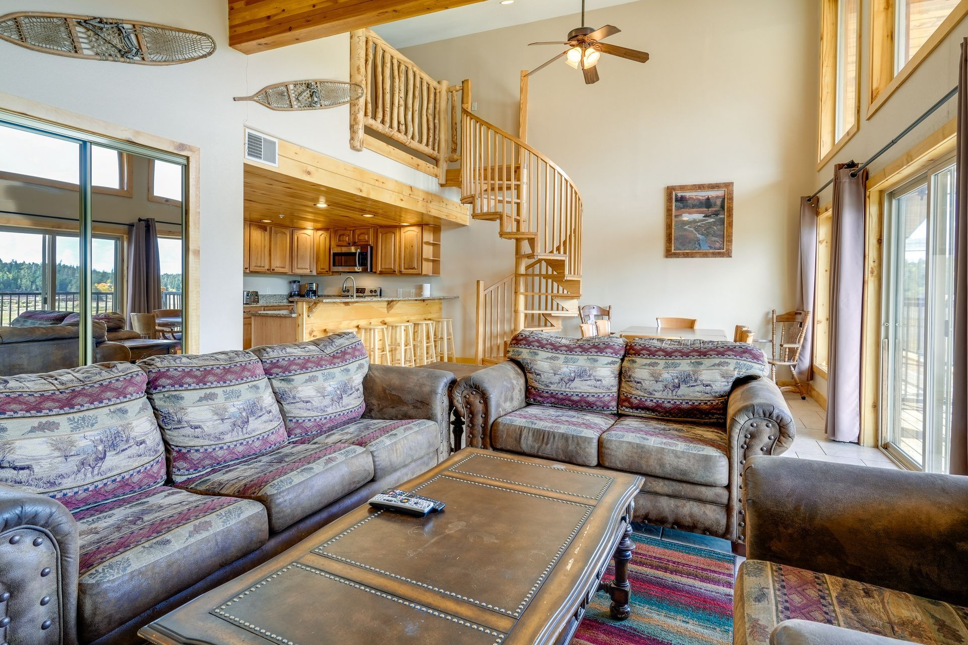 Spacious cabin living room with two floral-patterned sofas, wooden spiral staircase, and kitchen in the background.