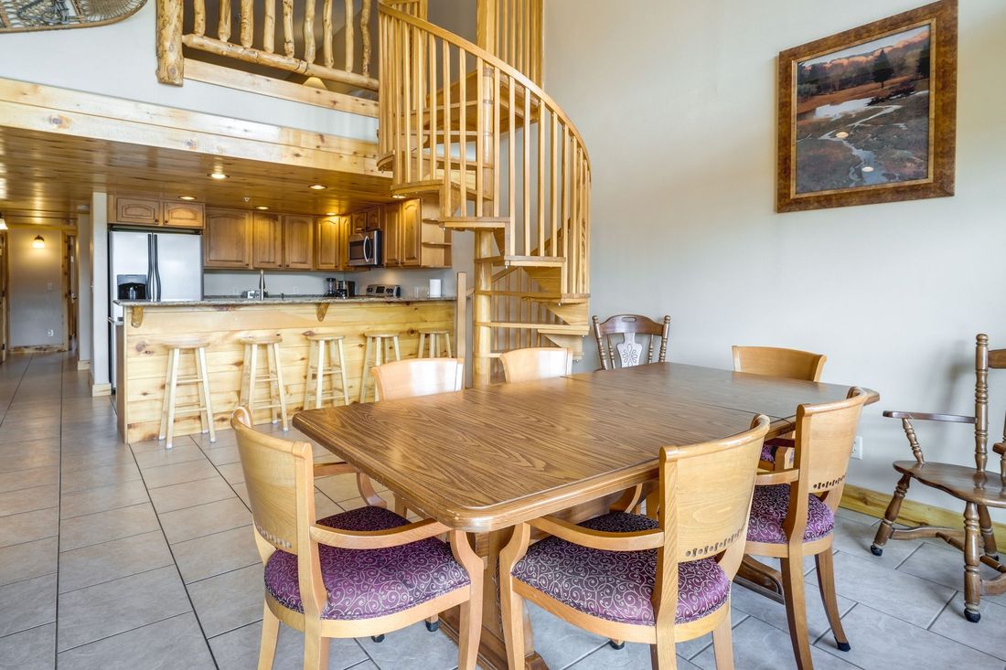 A dining area with a wooden spiral staircase, kitchen, and a table set for six.  The space is brightly lit.