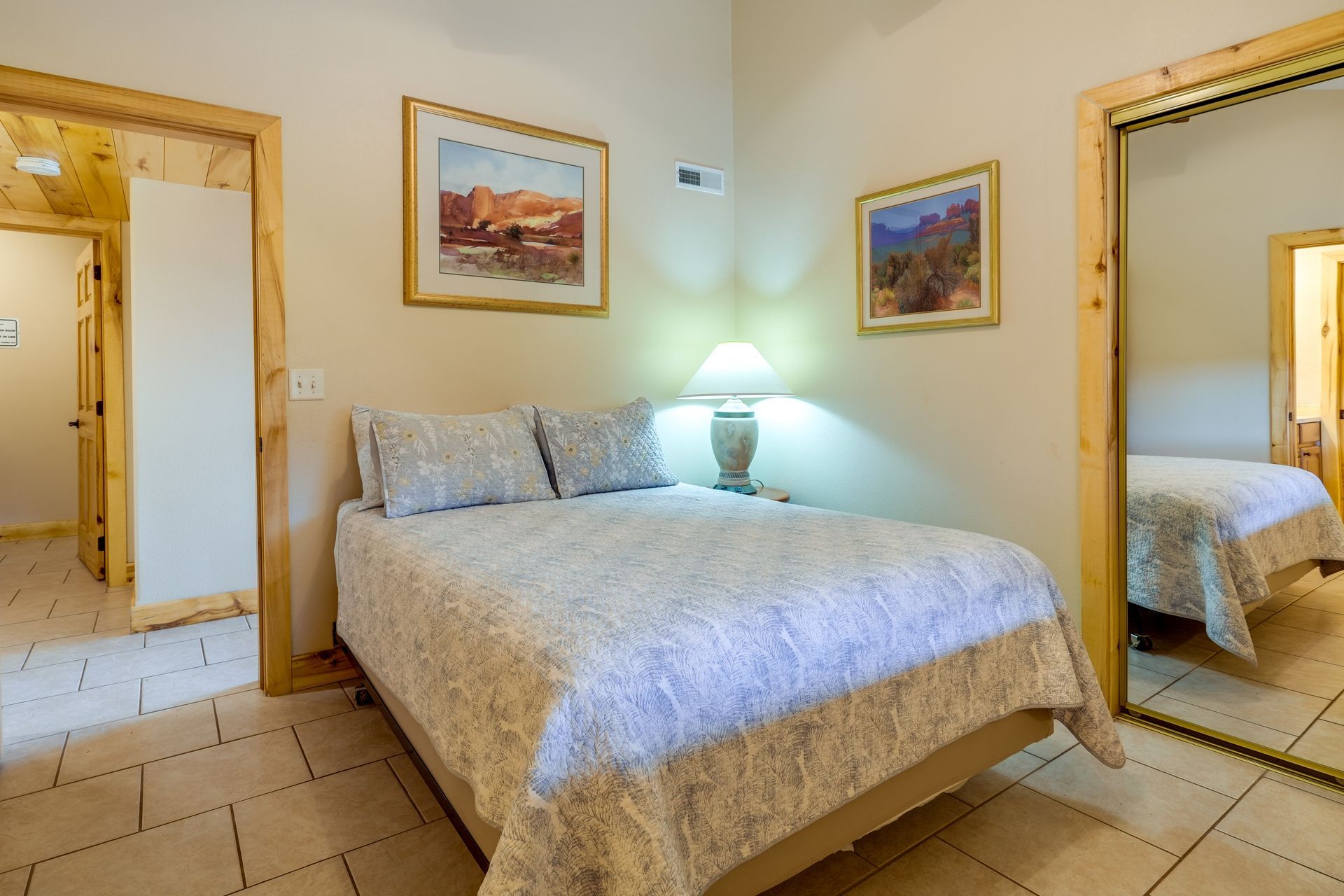 Bedroom with a double bed, two landscape paintings, and a tall mirror. Warm, neutral tones with tile flooring.