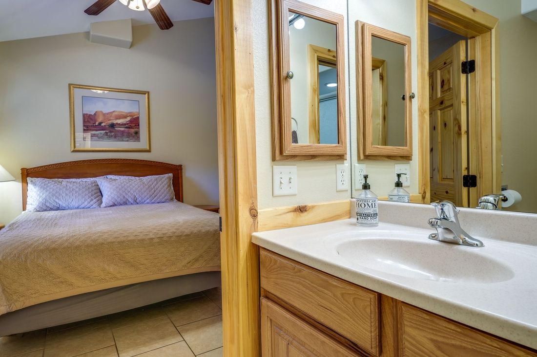 A bed with a wooden headboard is visible from the bathroom doorway. The bathroom has a sink and two mirrors.
