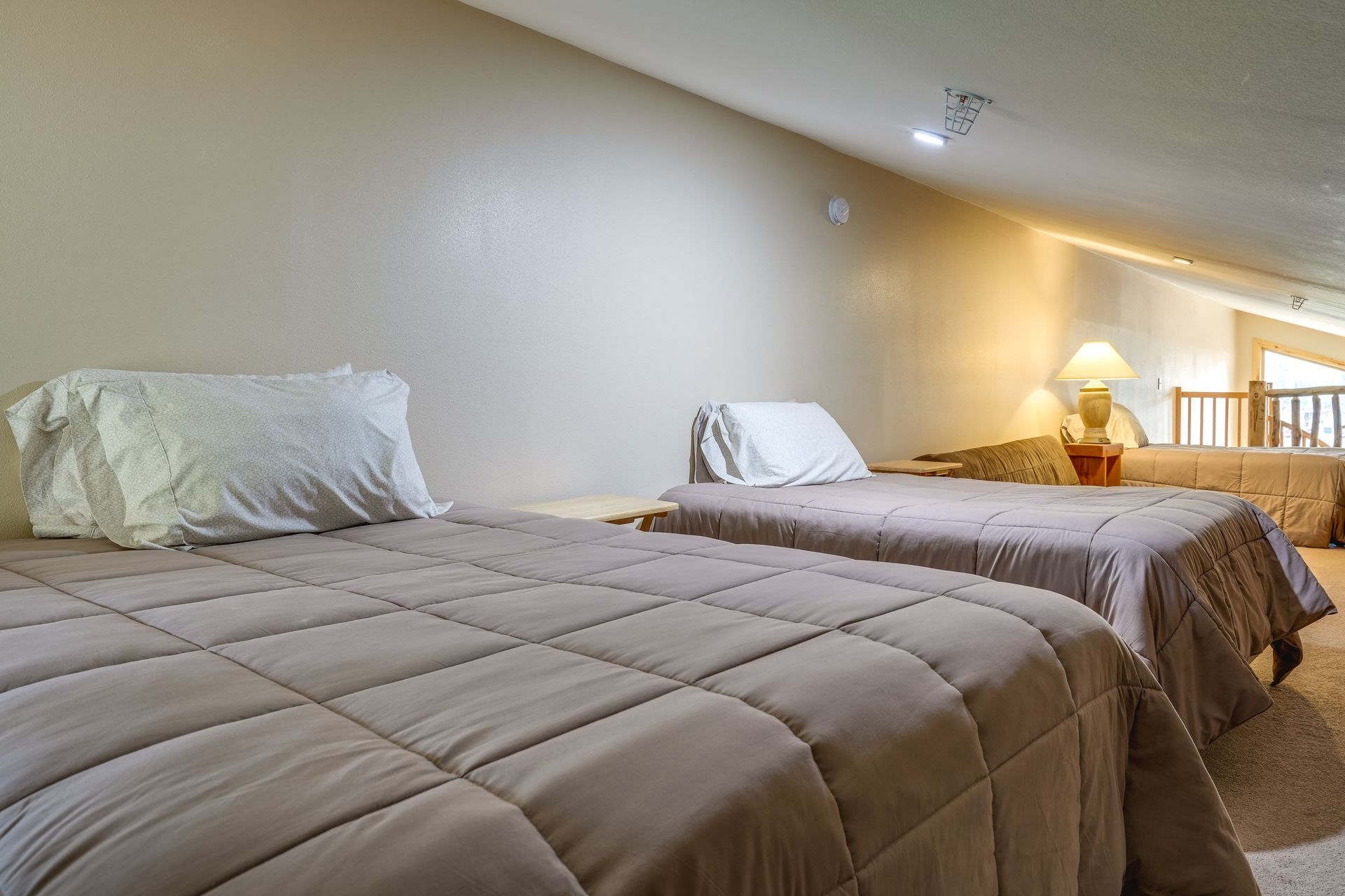 Two twin beds with beige quilts in a loft bedroom. A lamp and another bed are visible in the background.