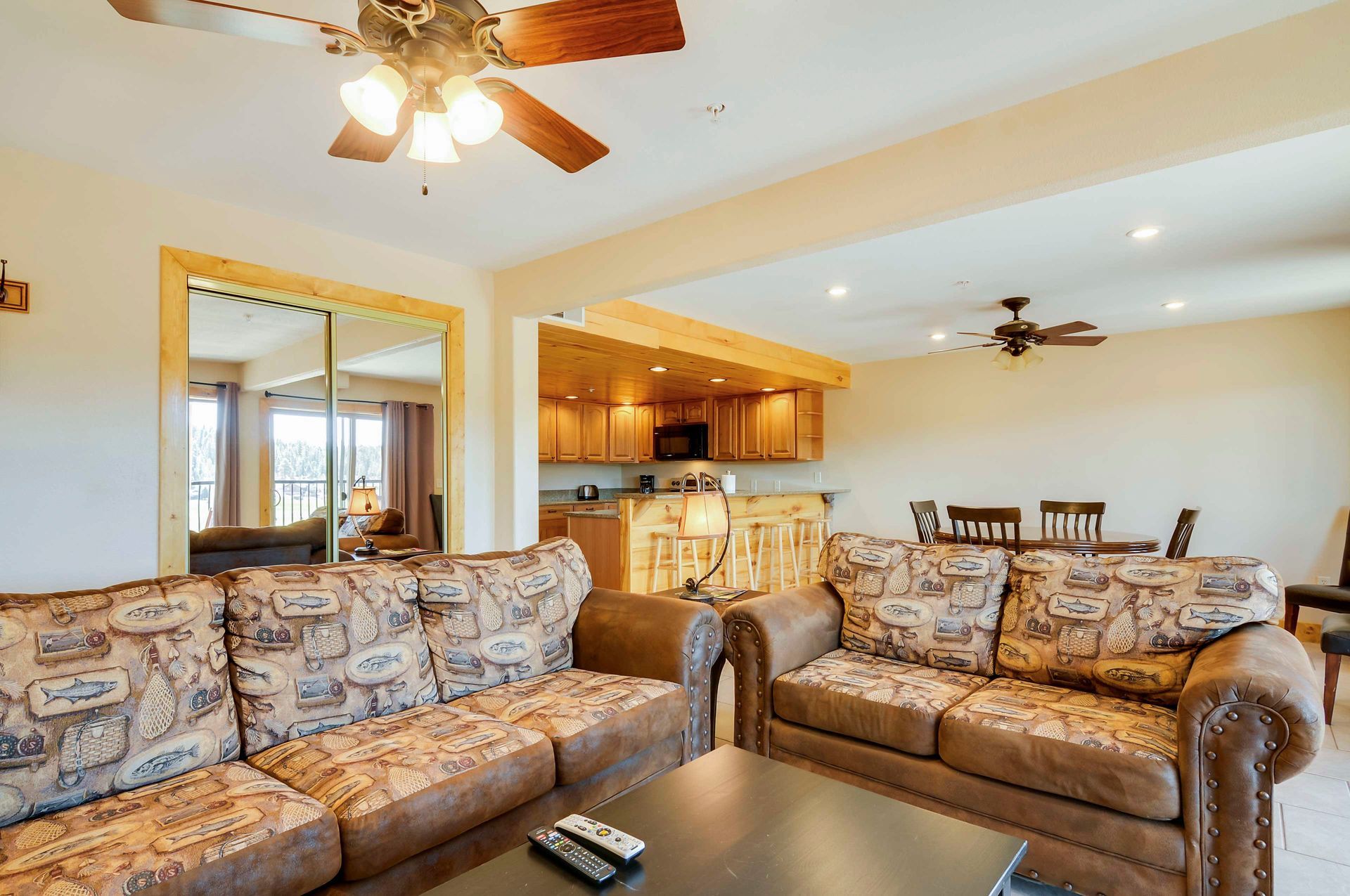 Spacious living room with two patterned sofas, a kitchen, and a dining area. Brown and beige tones with a ceiling fan.