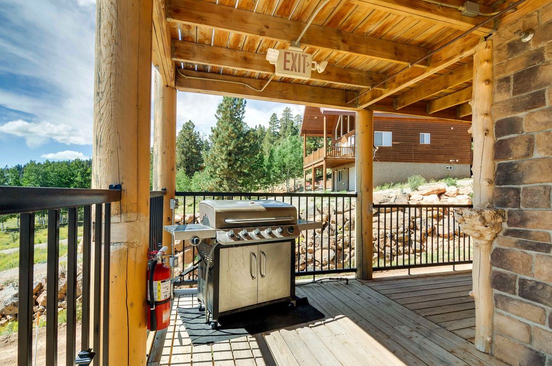 Outdoor wooden deck with a grill and a fire extinguisher. The view includes trees, another building, and a blue sky.