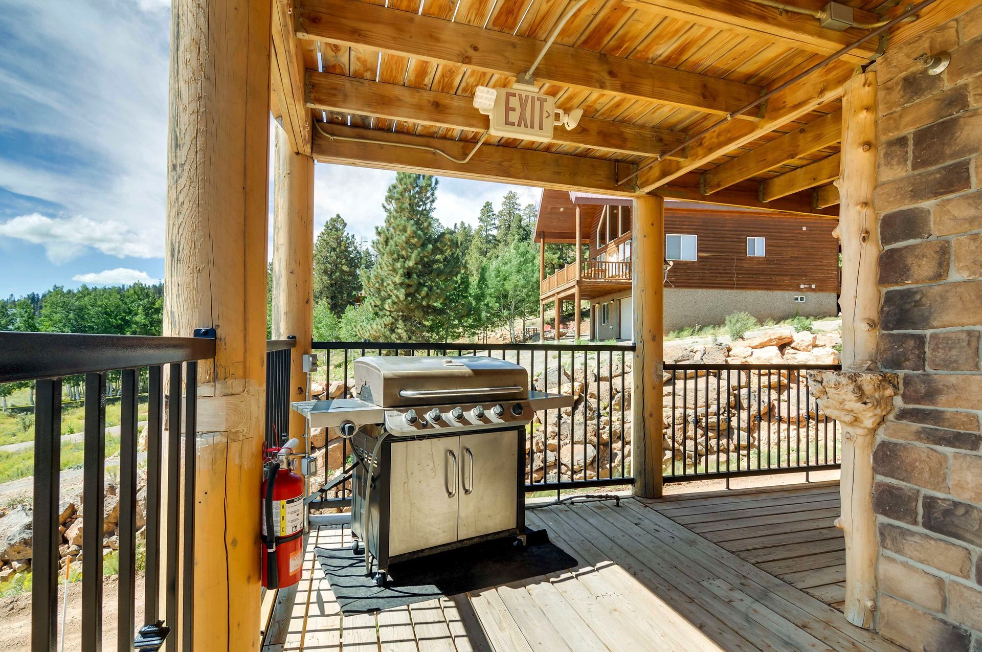 A covered outdoor patio with a grill, fire extinguisher, and view of trees and another house.