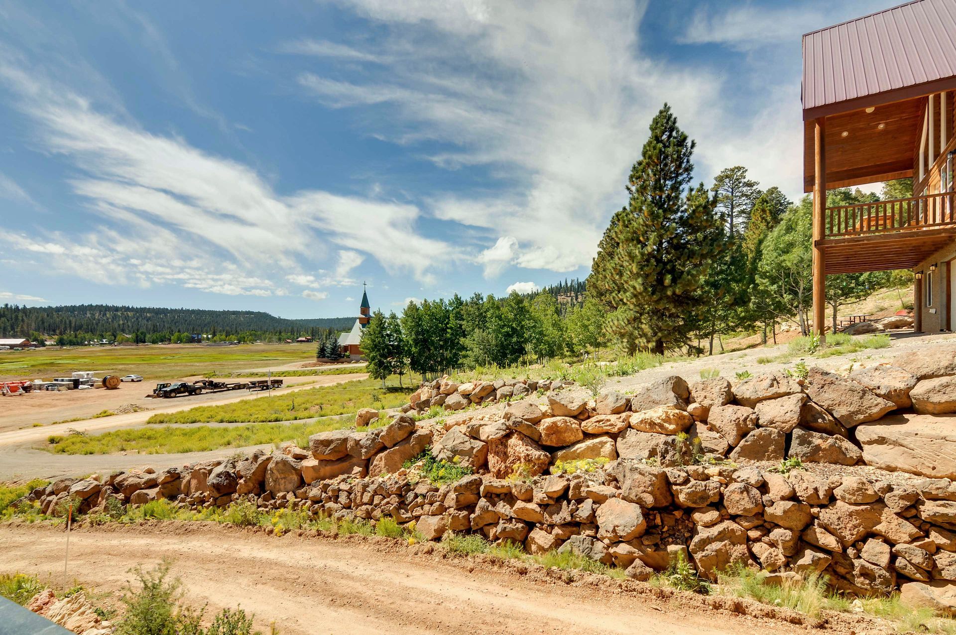 Dirt road leads past a rock wall towards a cabin with a balcony under a bright blue sky and scattered clouds.