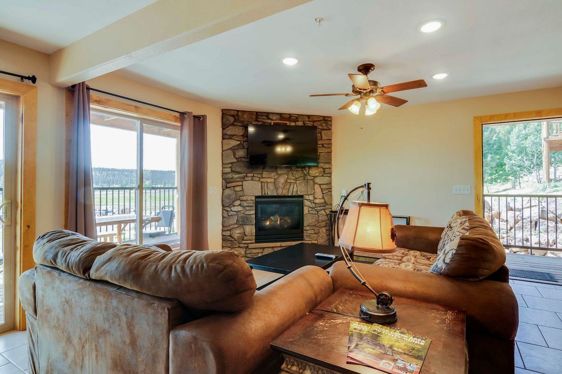 Cozy living room with a stone fireplace, TV, brown couches, and a view of a balcony and surrounding greenery.