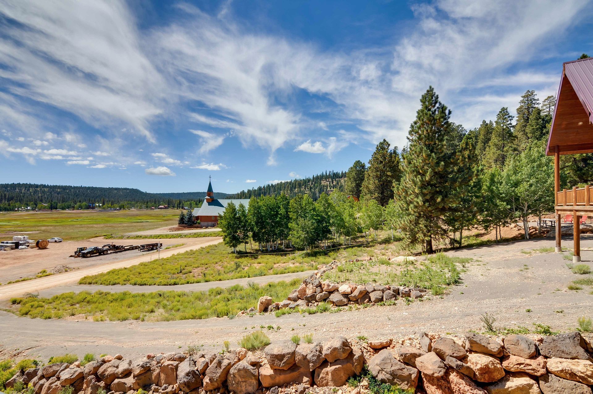View of a mountain landscape with a stone wall in the foreground, a dirt road, trees, and a building in the distance.