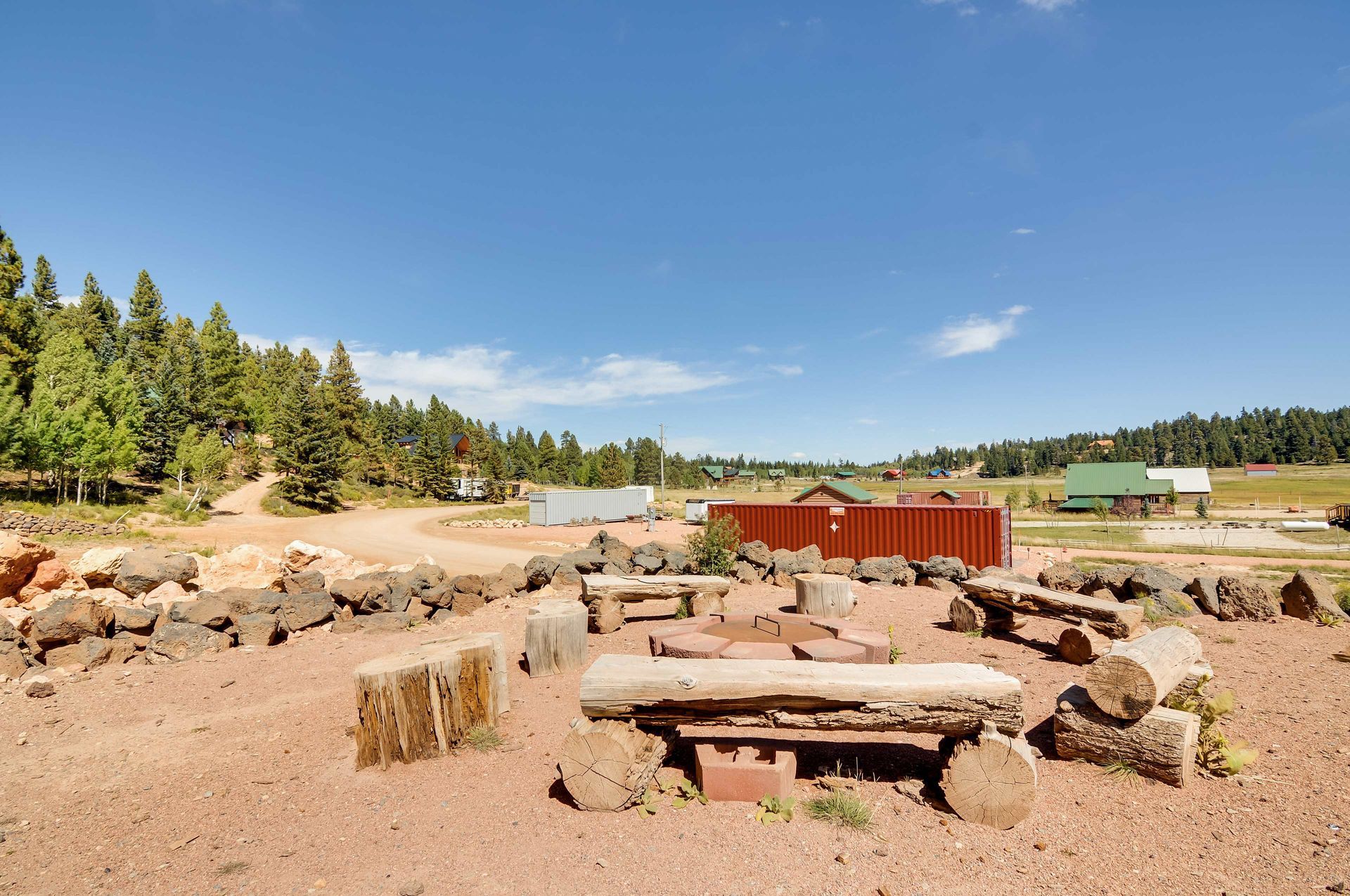 A fire pit area with log benches, surrounded by reddish dirt, under a blue sky. Trees line the sides of the space.