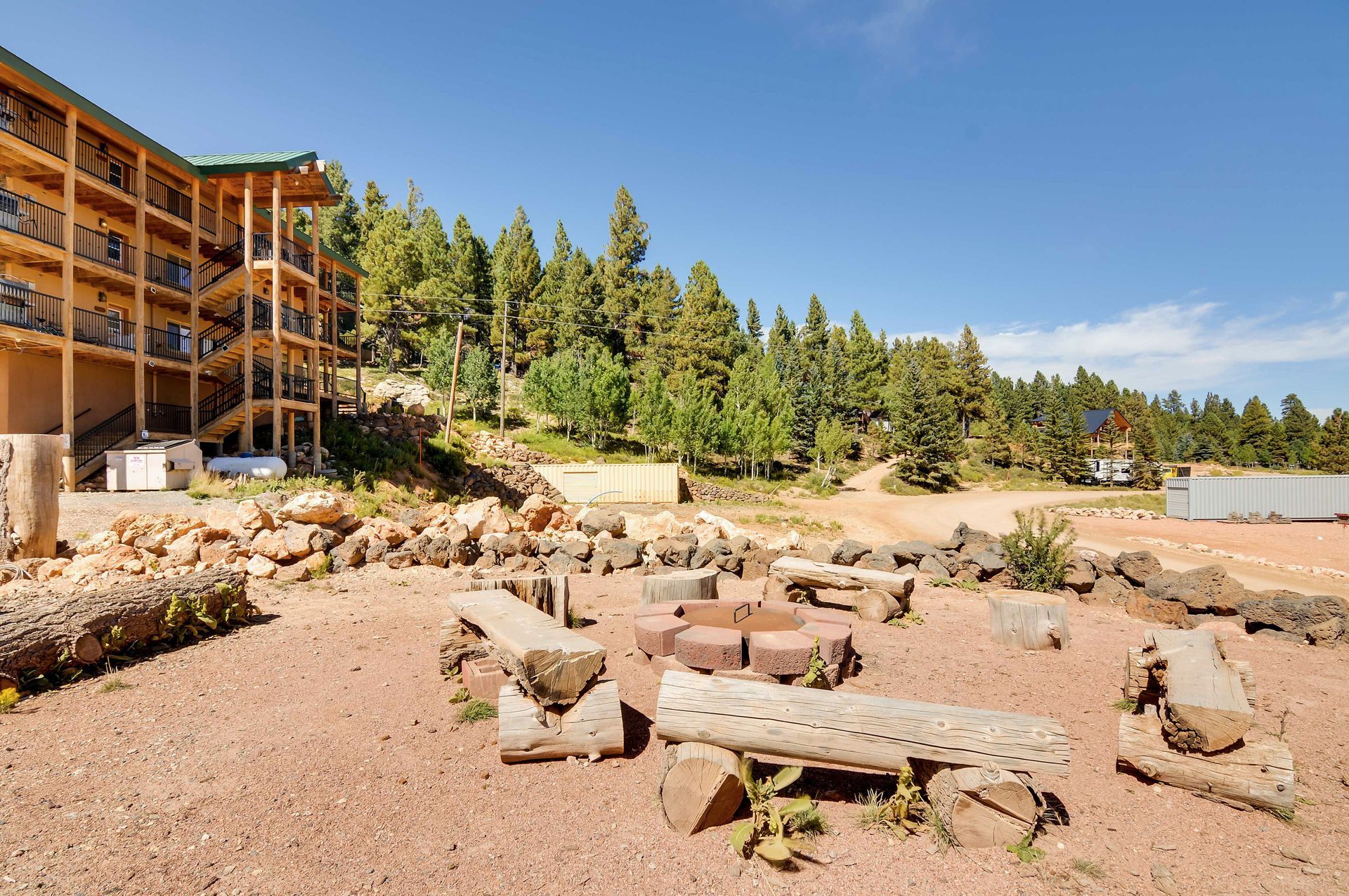 A fire pit with log benches in front of a multi-story building and pine trees under a blue sky.