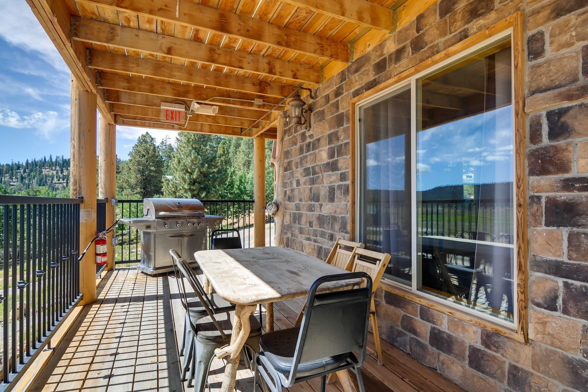 Outdoor patio with a wooden table and chairs, grill, and a view of trees and a mountain under a blue sky.