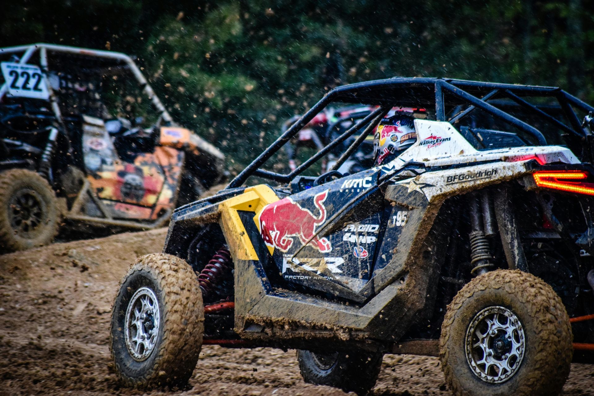 Two mud-covered UTVs racing on a dirt track. The lead vehicle is branded with Red Bull logos.