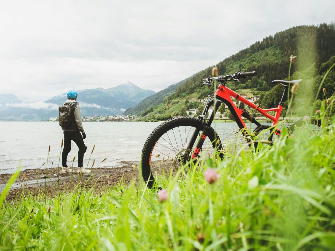 Person standing in water, looking at a lake and mountains, with a red mountain bike on the grassy bank. Overcast day.