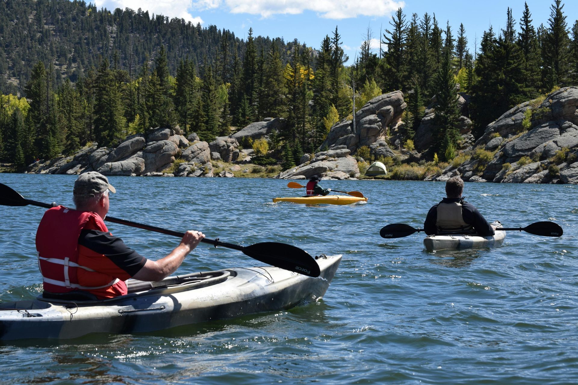 Three people kayaking on a lake, surrounded by trees and rocks under a blue sky. One wears a red life vest.