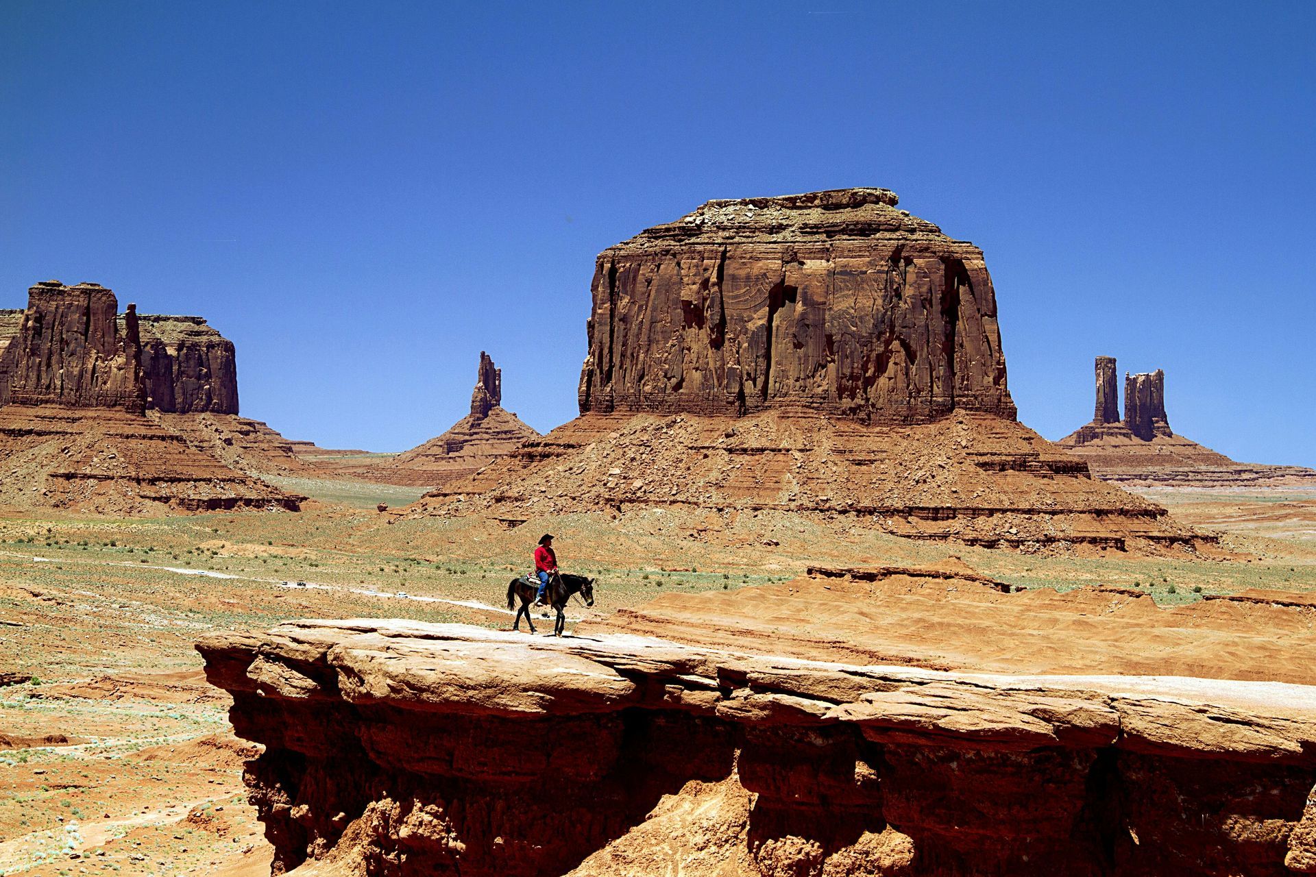 Cowboy on horseback surveys the vast desert landscape with iconic red rock formations under a bright blue sky.