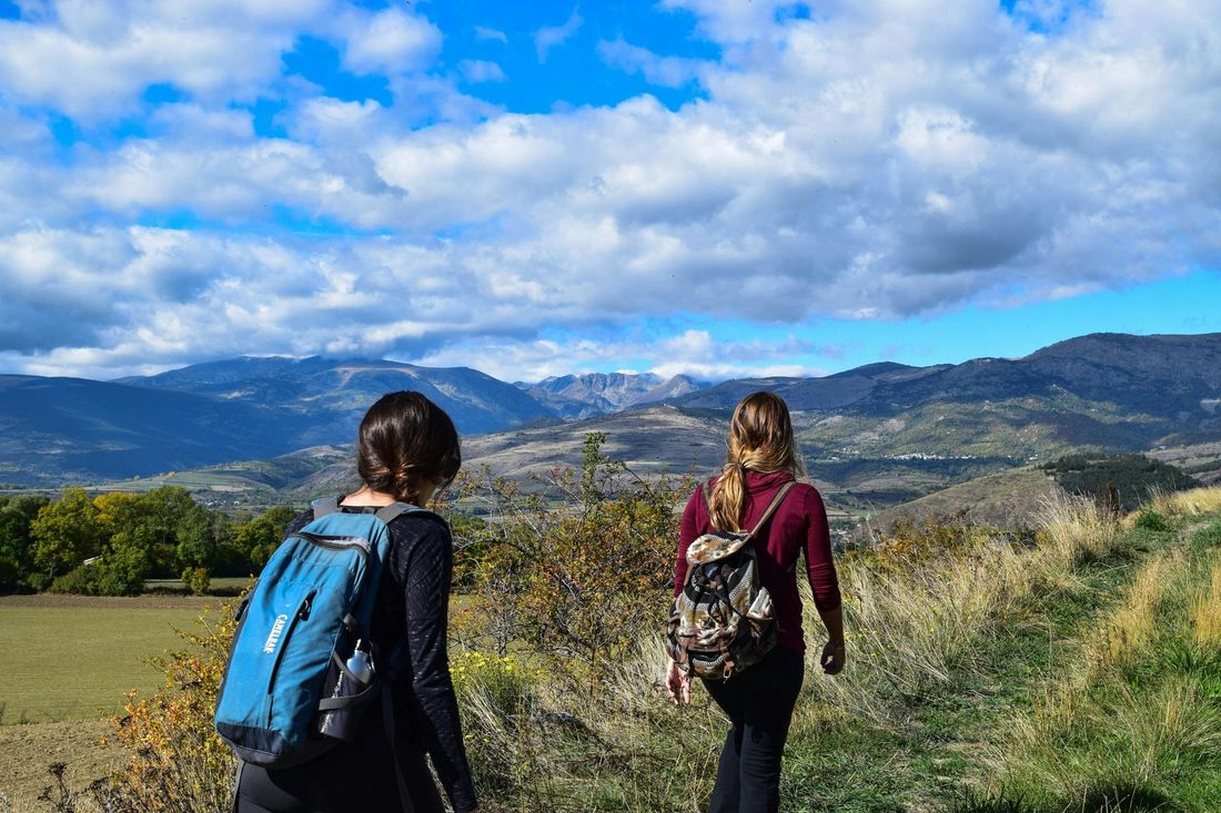 Two people hike on a trail with backpacks, overlooking mountains under a cloudy blue sky.