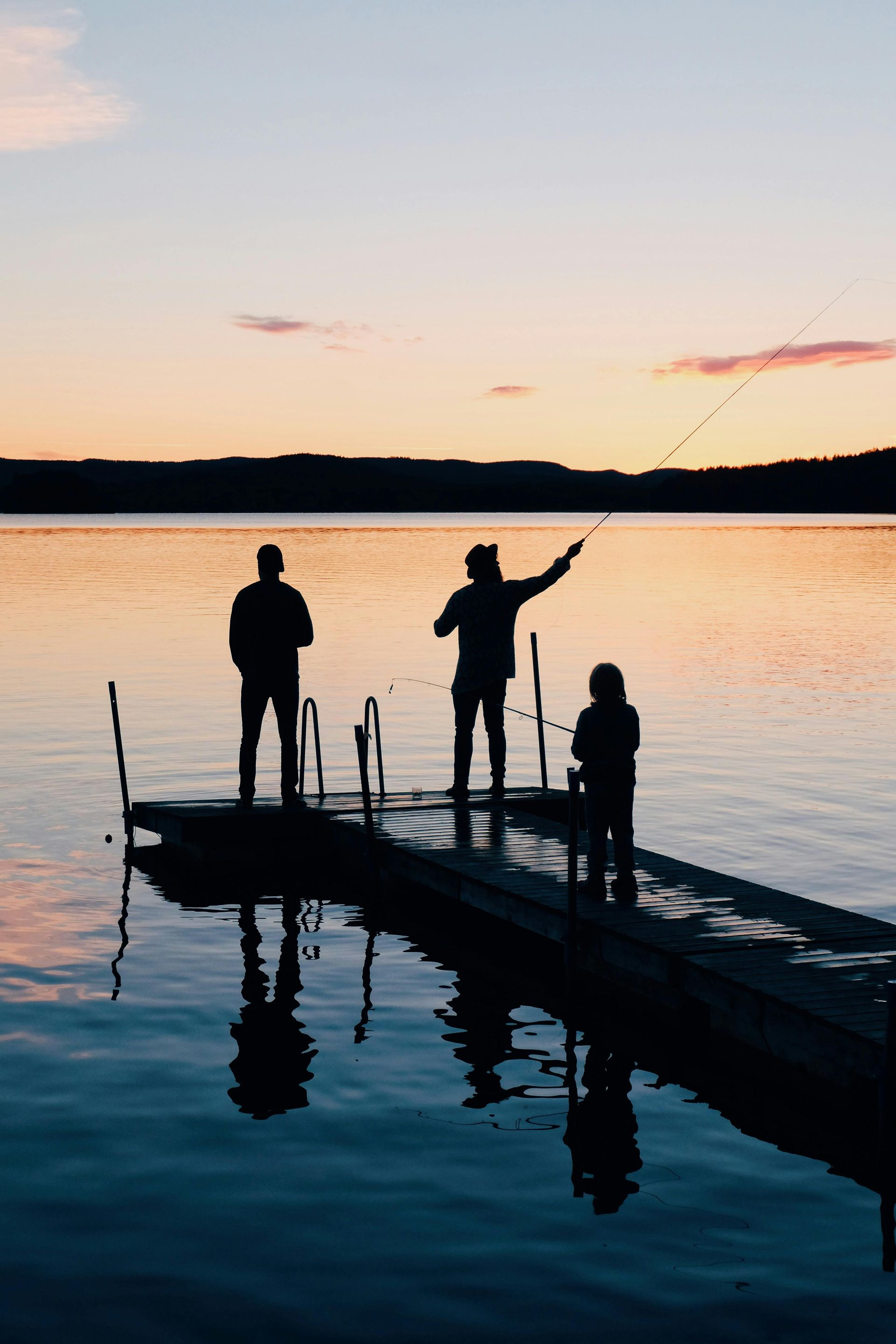 Silhouetted figures fishing off a dock at sunset over calm water; soft orange and blue hues.