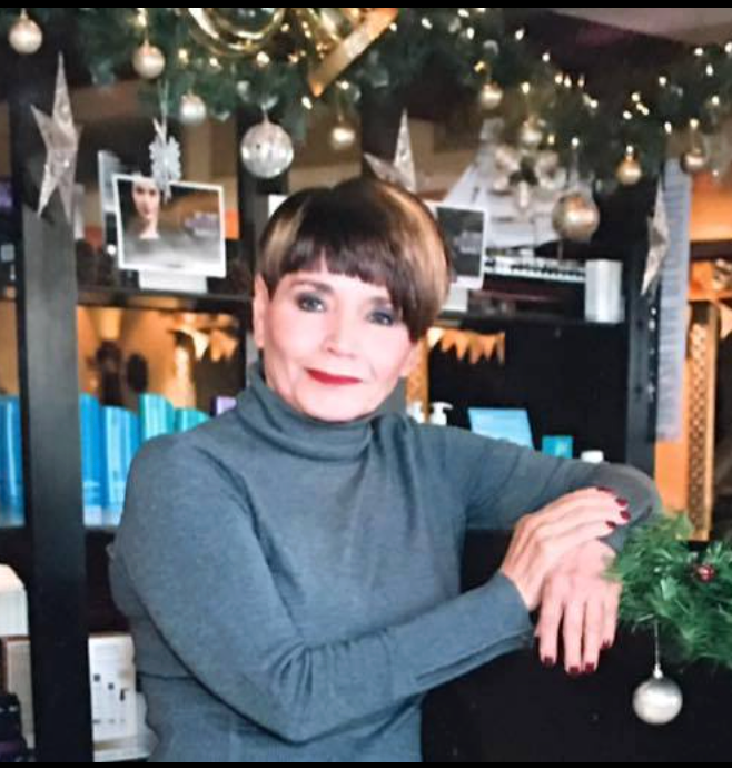 Woman with short, dark hair, wearing a gray turtleneck, leaning on a counter, with Christmas decorations in the background.