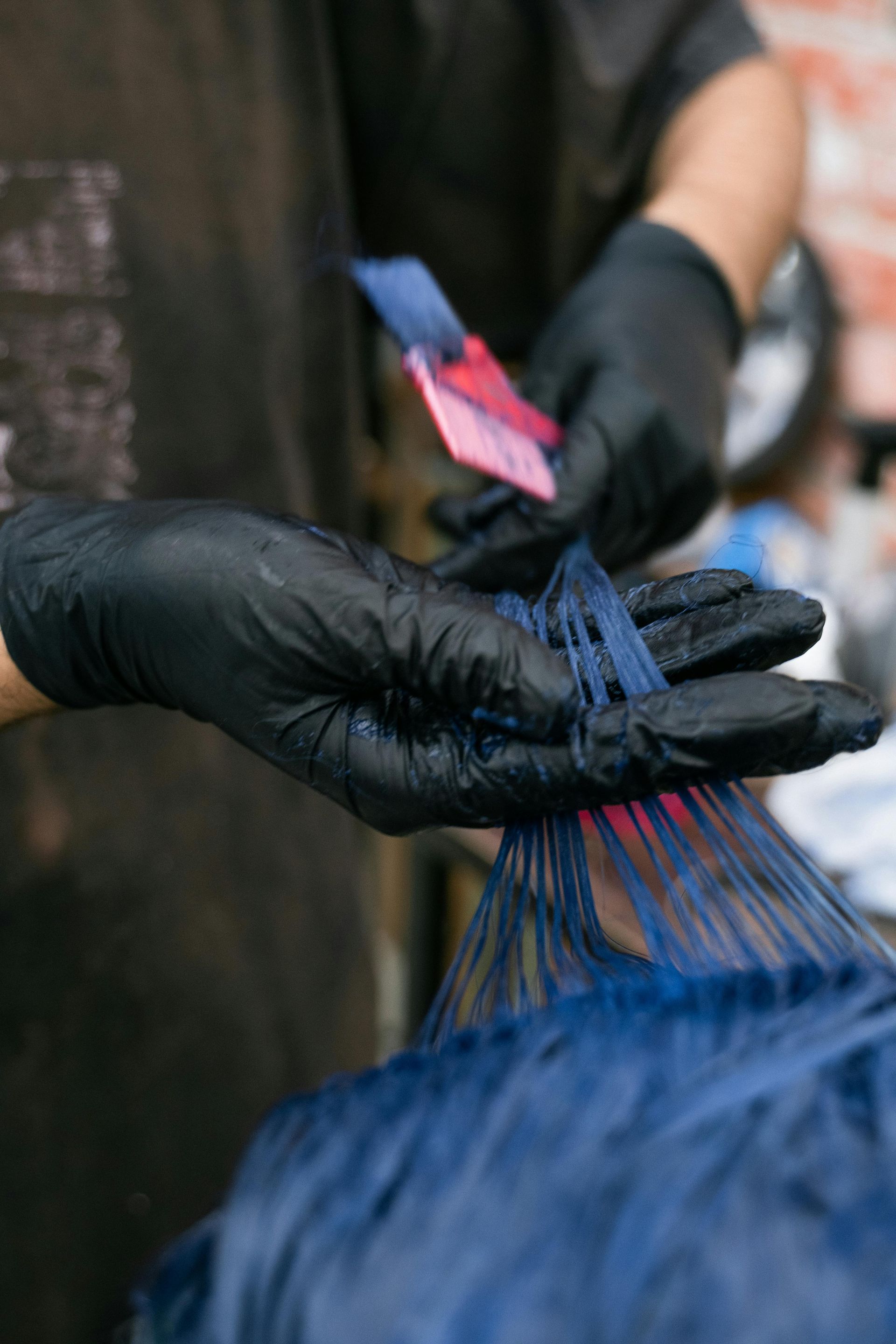 Hands wearing black gloves dyeing blue hair with a color swatch in a salon.