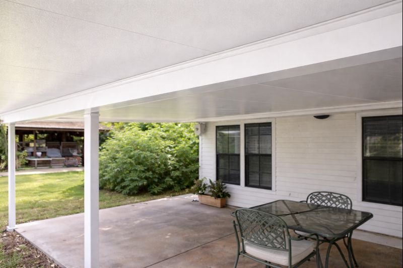 Patio with white roof, concrete floor, table, and chairs. Green foliage and white house.