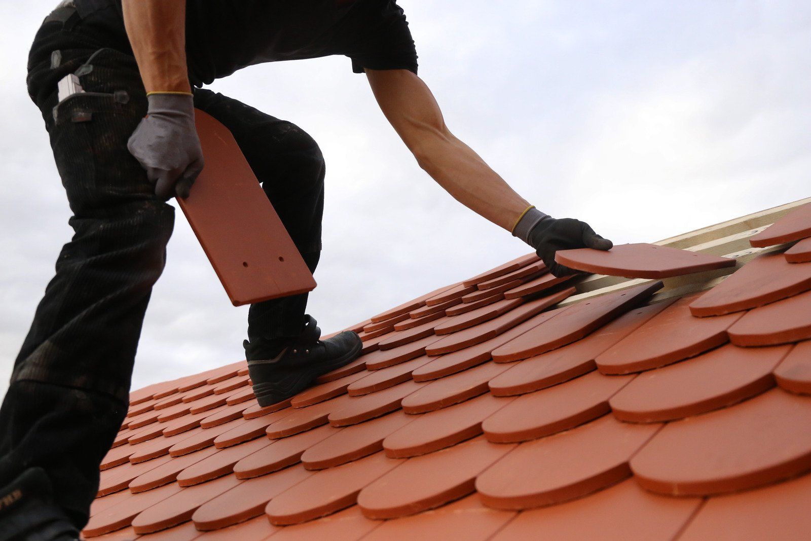 Roofer installing red tile roof. Roofer wears black gloves and pants. Overcast sky in background.