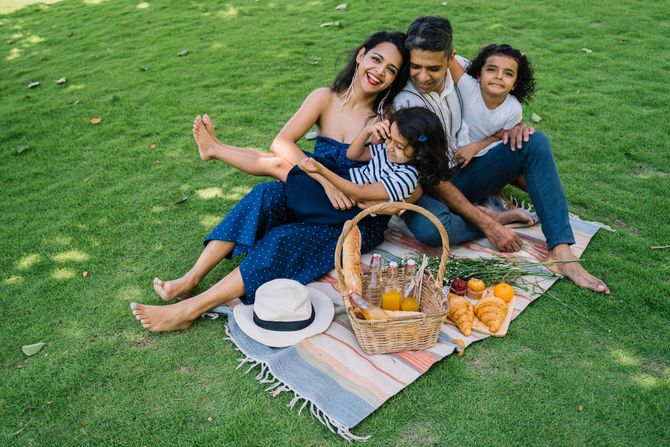A family of four sitting on a picnic blanket on green grass, smiling and interacting with a picnic basket.