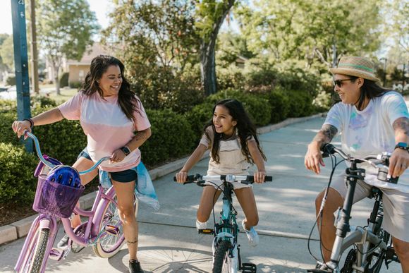 Three people smiling while riding bicycles along a sunny, tree-lined path.