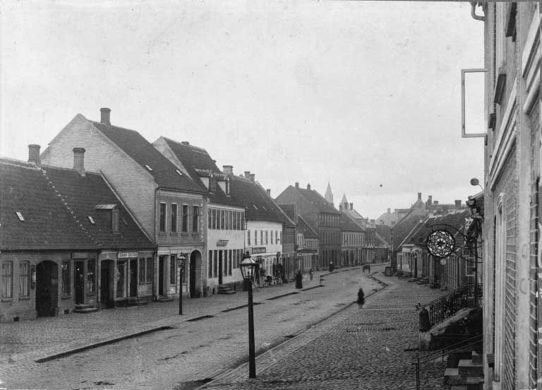 Sort-hvid gadescene med rækkehuse, brosten og et par fodgængere på en stille, gammel byvej