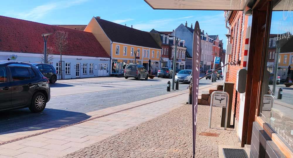 Solrig gade med parkerede biler, murstensbygninger og butikker i en lille by.