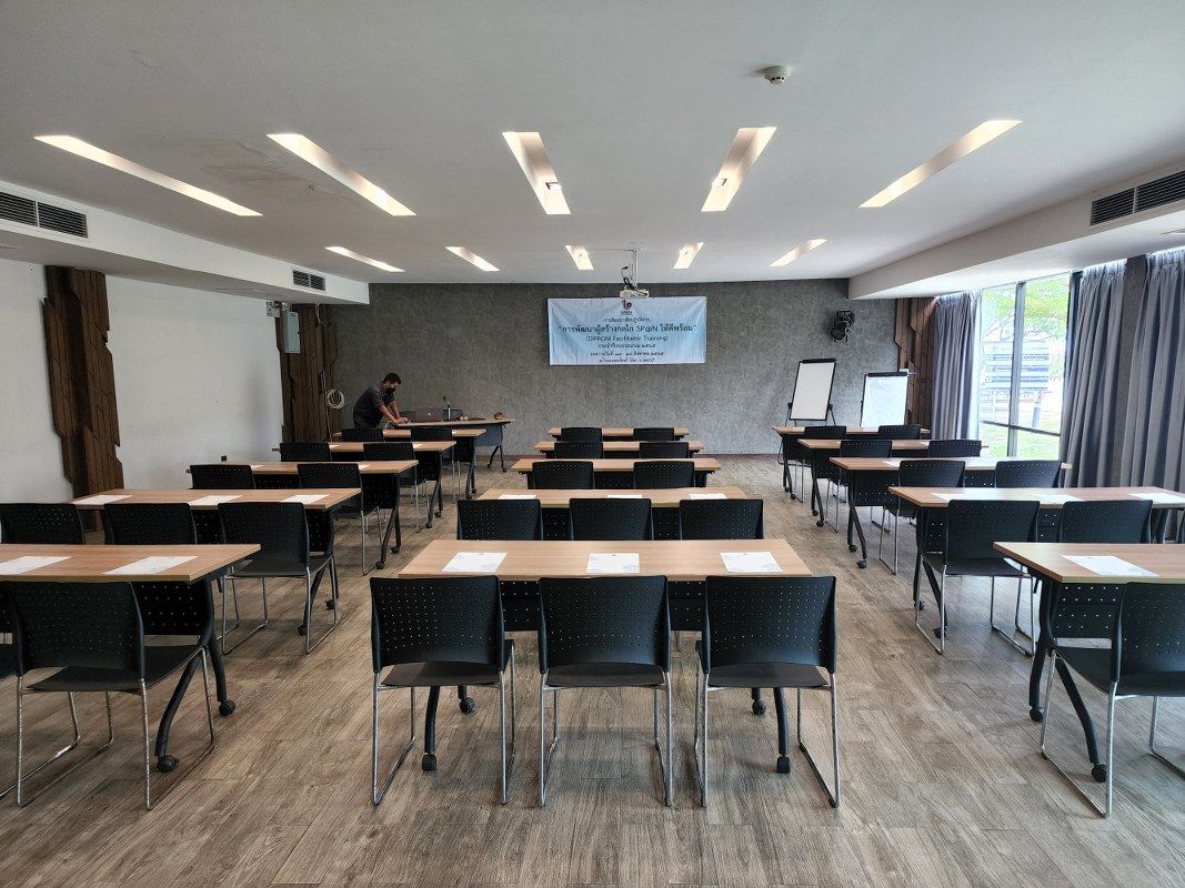 Classroom with rows of desks, chairs, and a person working at a desk. Projector on the ceiling.