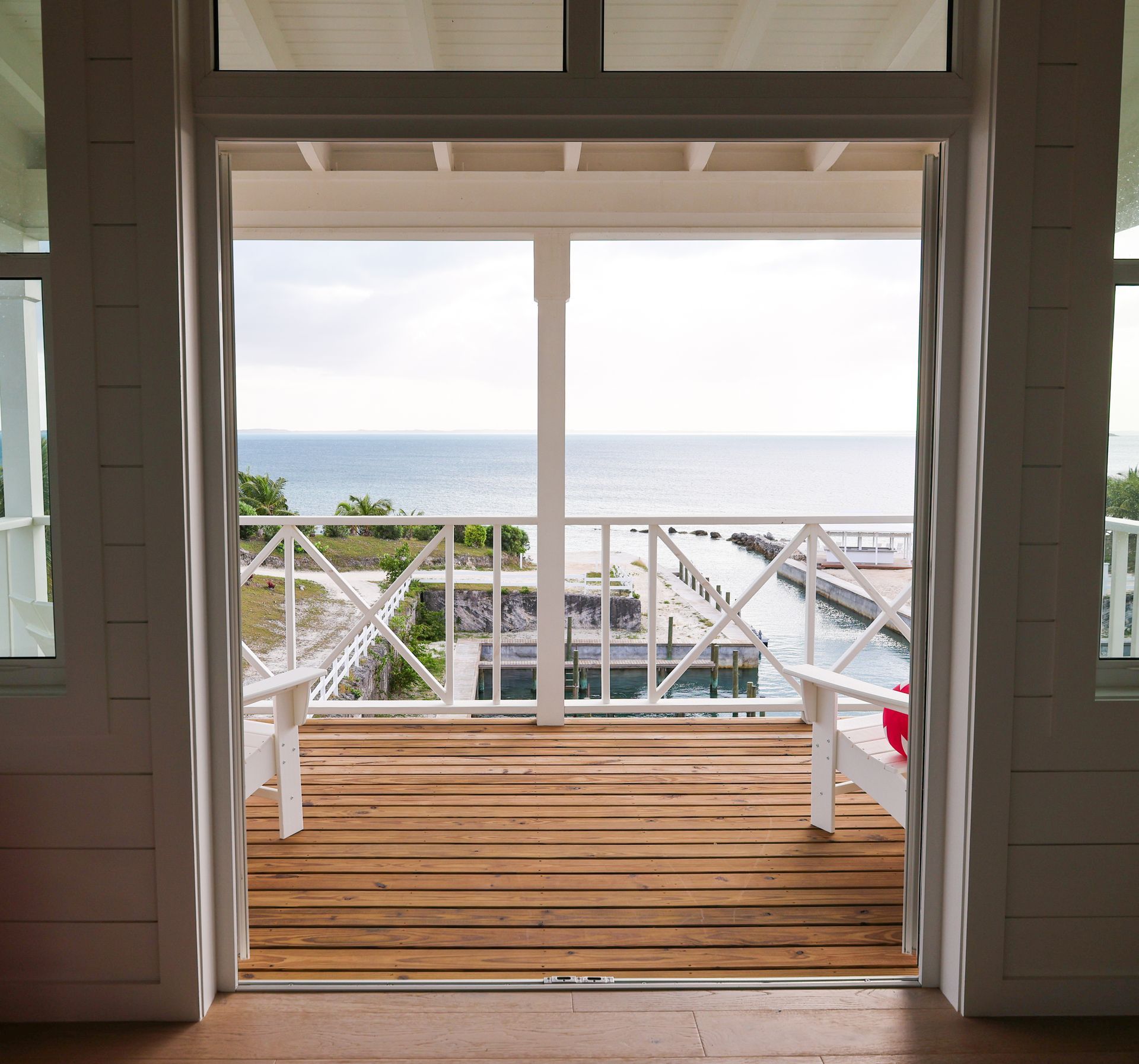 Balcony view of ocean and dock. White wooden railing, deck, and chairs. Cloudy sky.