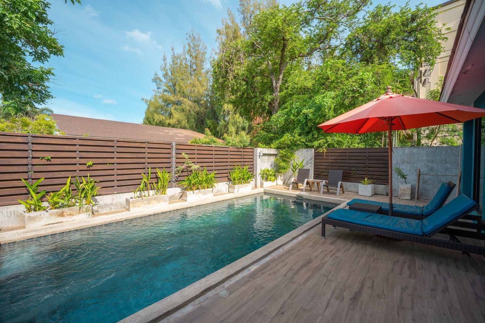 Swimming pool with lounge chairs and a red umbrella, surrounded by trees and a wooden fence.