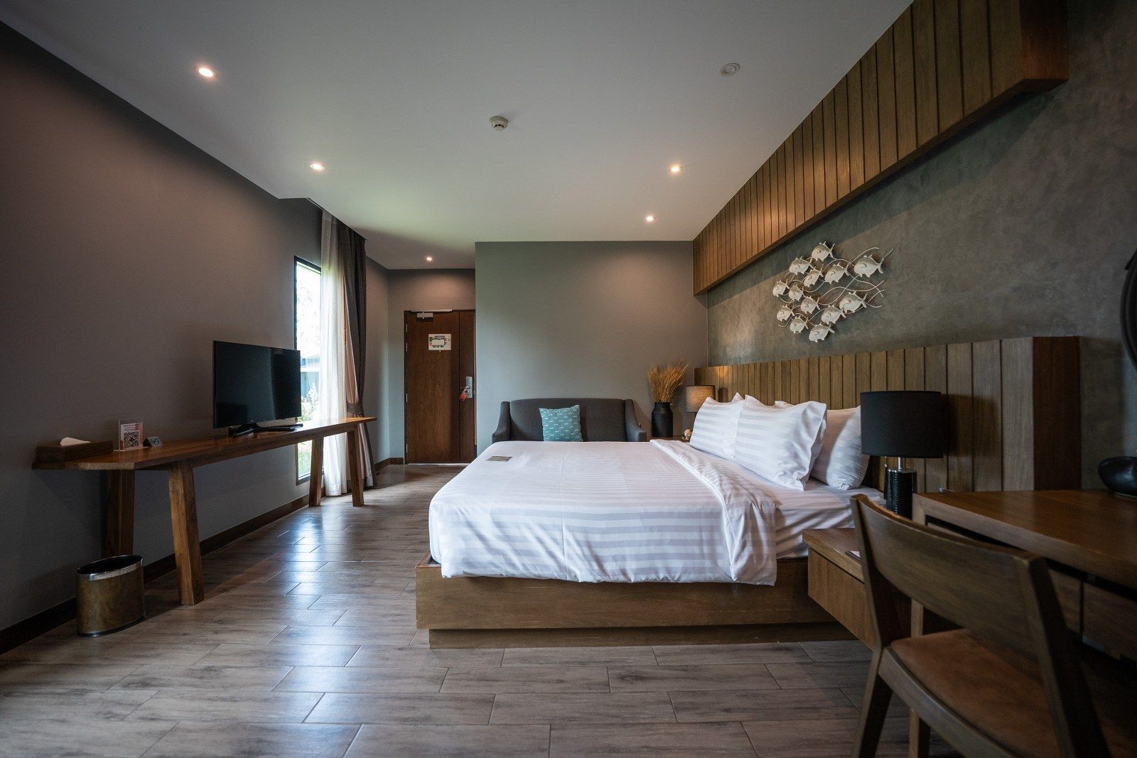 Hotel room with bed, sofa, TV, wooden desk, and decorative wall art. Gray walls and wood flooring.