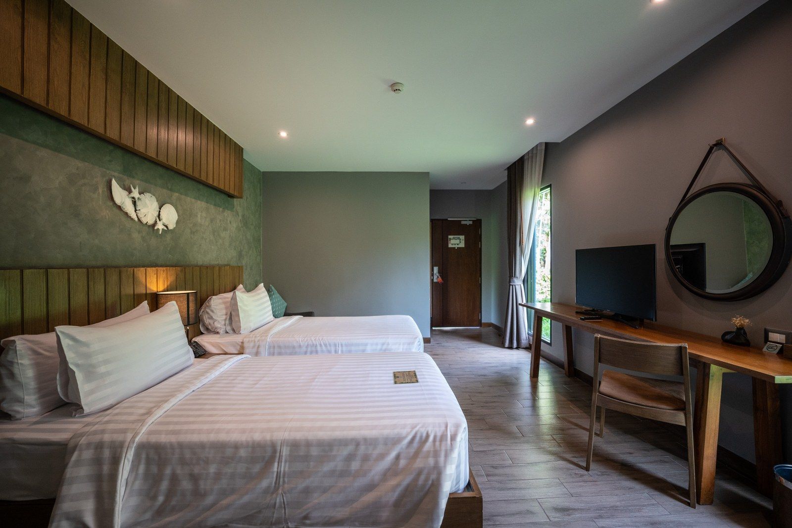 Hotel room with two beds, wooden desk, mirror, and television; gray walls, wood trim.
