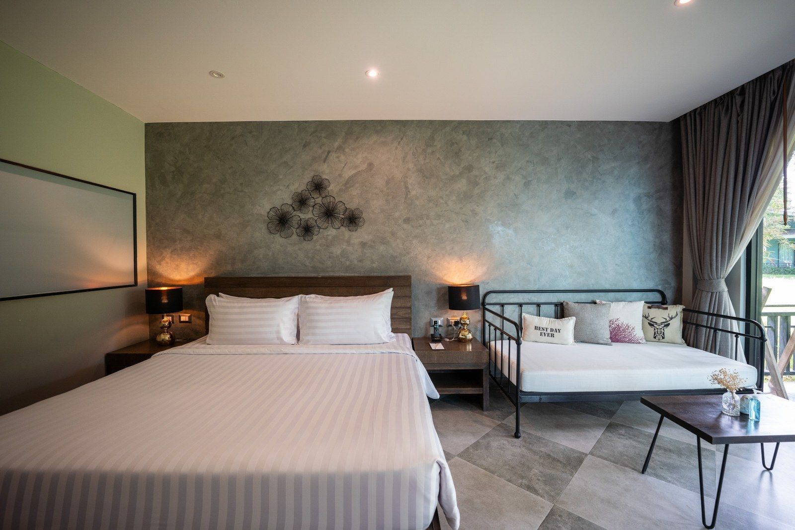 Bedroom with a bed, a daybed, and a small table; a decorative wall piece and a view through the curtains.