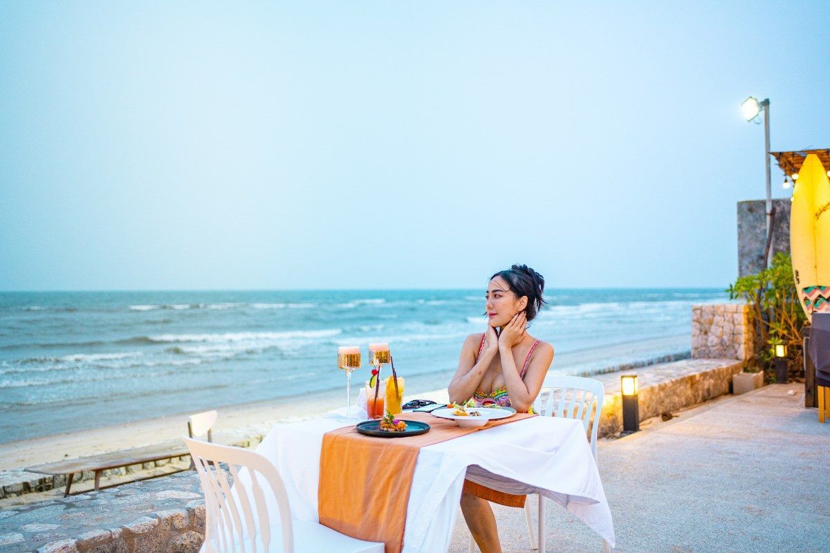 Woman sits at table set for dinner by the beach, gazing at the ocean.