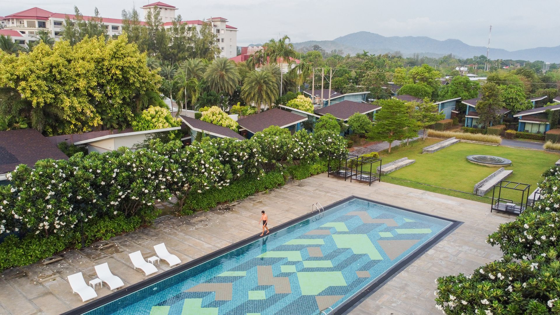Aerial view of resort with a pool, buildings, and greenery. A person stands in the pool.