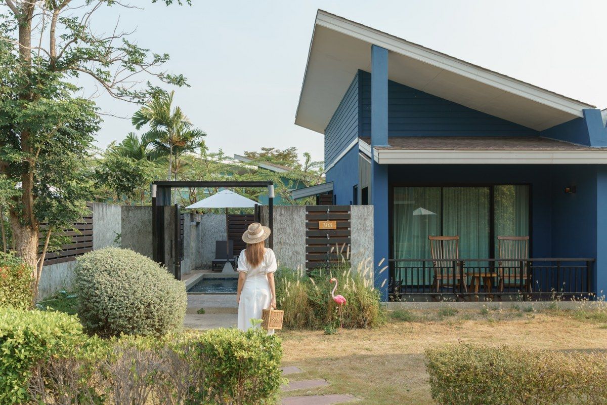 Woman in white dress walks toward blue house with patio, carrying a basket.