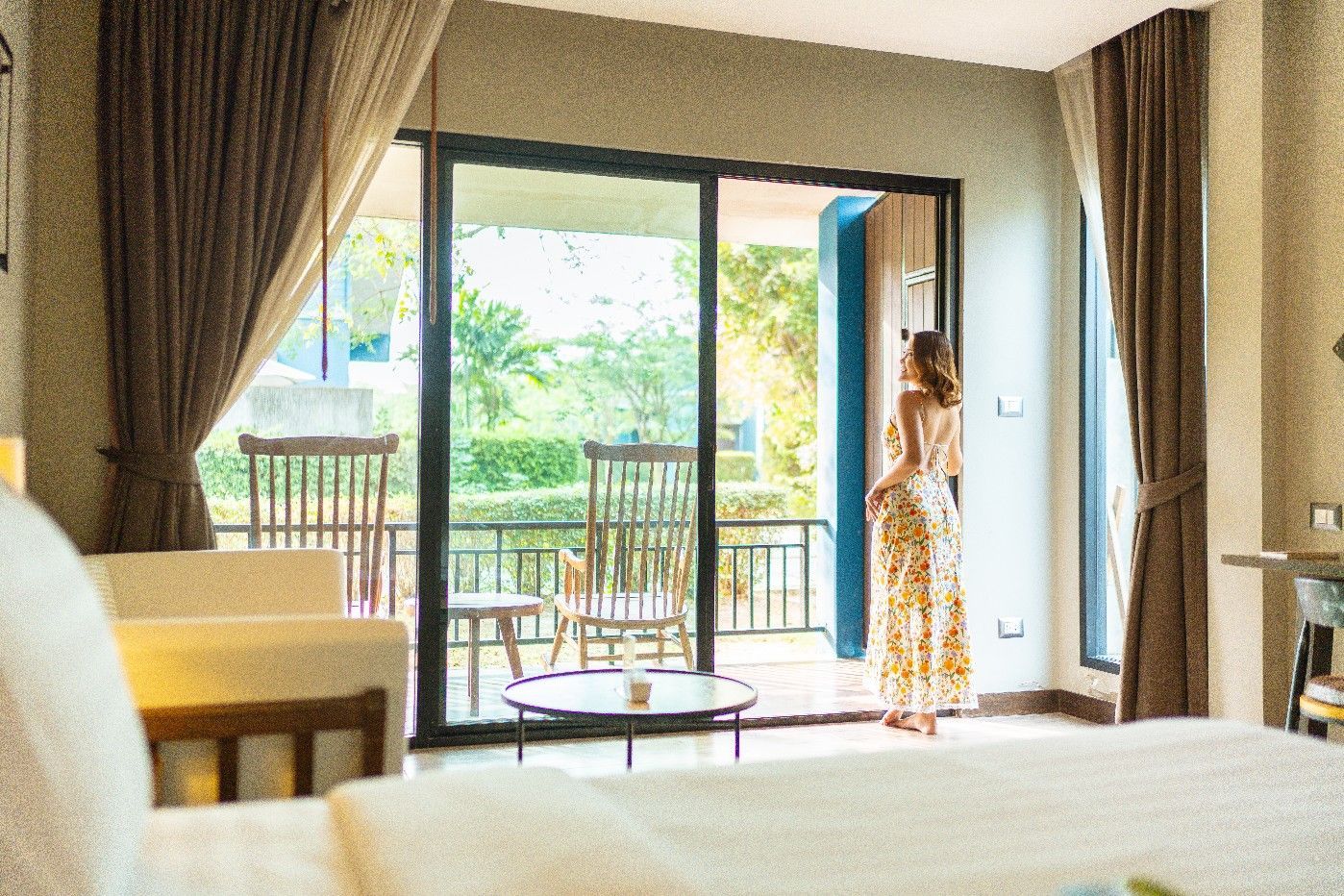 Woman in sundress stands at balcony door, looking out at trees.
