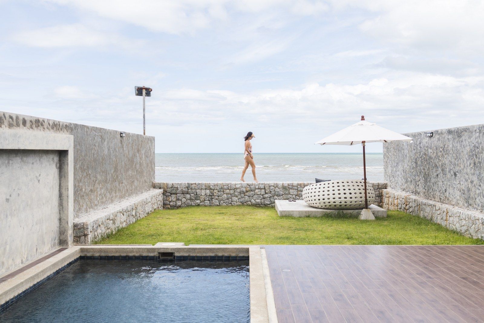 A person walks along a wall by the beach. A pool and lounge area in foreground. Cloudy sky.