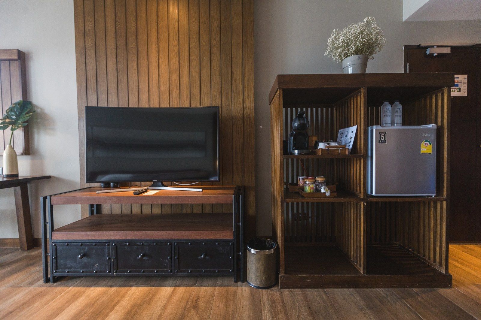 Wooden TV stand, open shelves, mini fridge, flower pot in a hotel room with wood paneling.