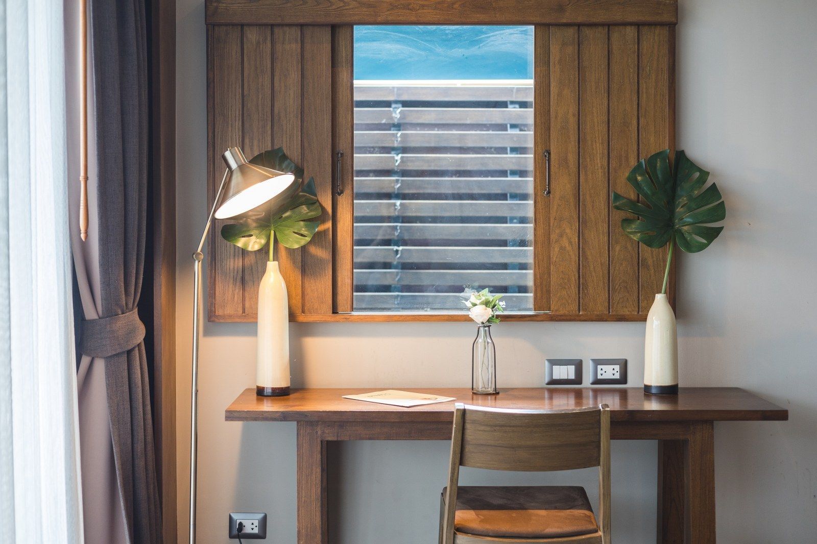 Wooden desk by a window with Monstera leaves and a chair.