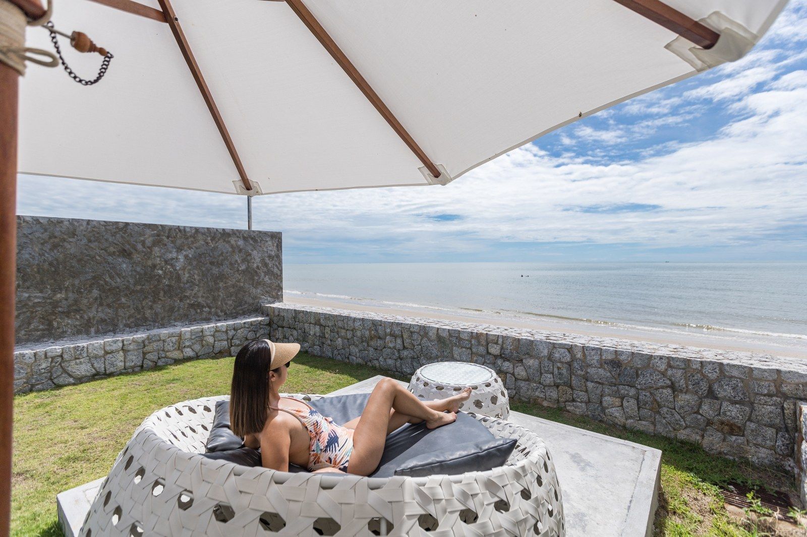 Woman relaxing on a white woven chair under an umbrella, near a beach.