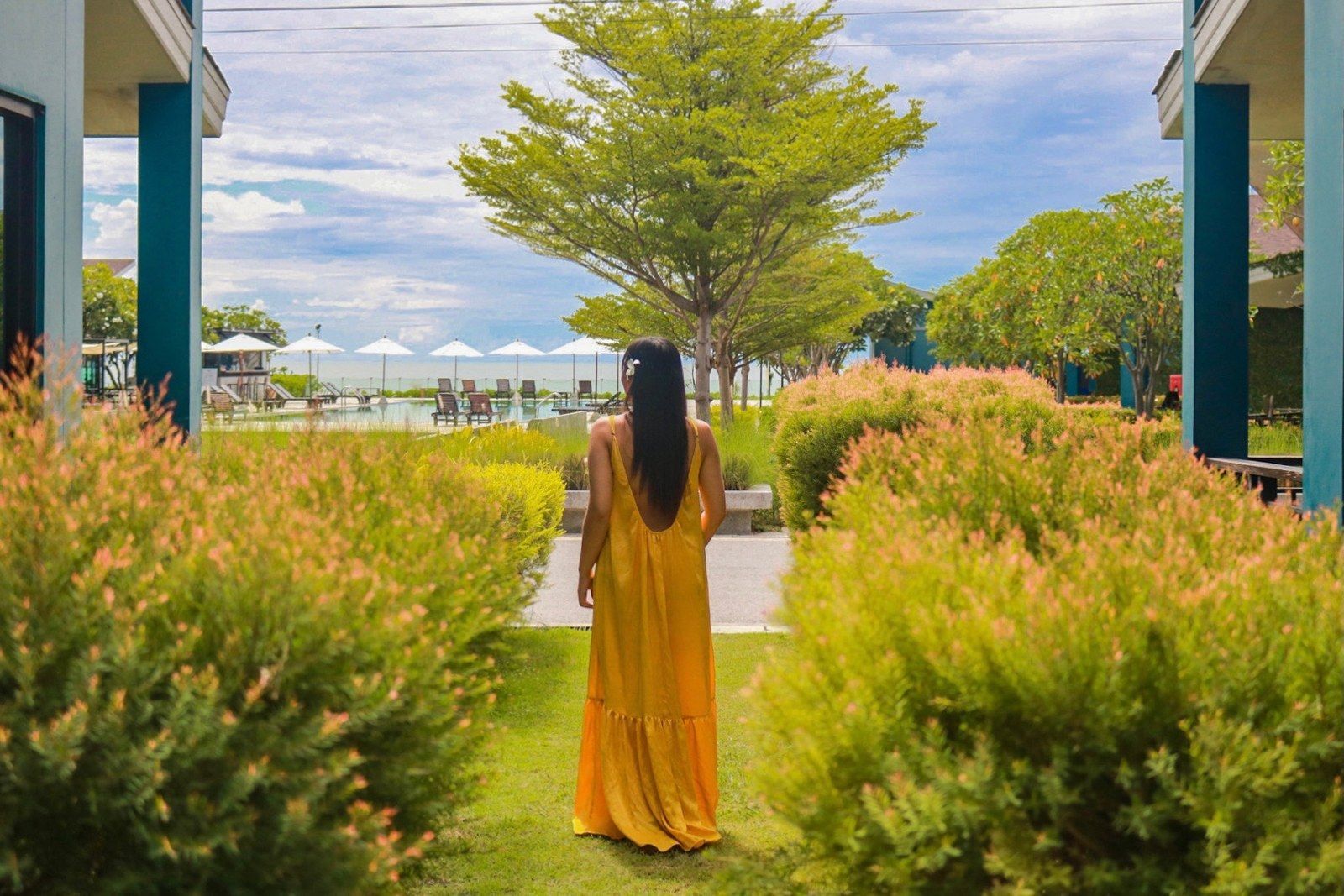 Woman in yellow dress walking toward ocean view between hedges and buildings.