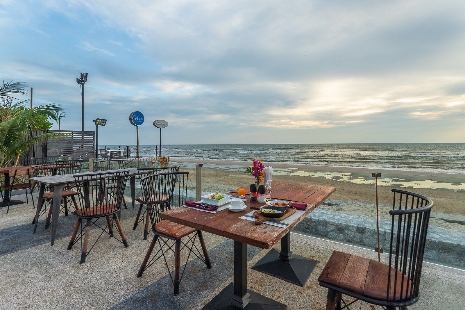 Outdoor event setup near the beach with round tables, buffet, and buildings under a cloudy sky.