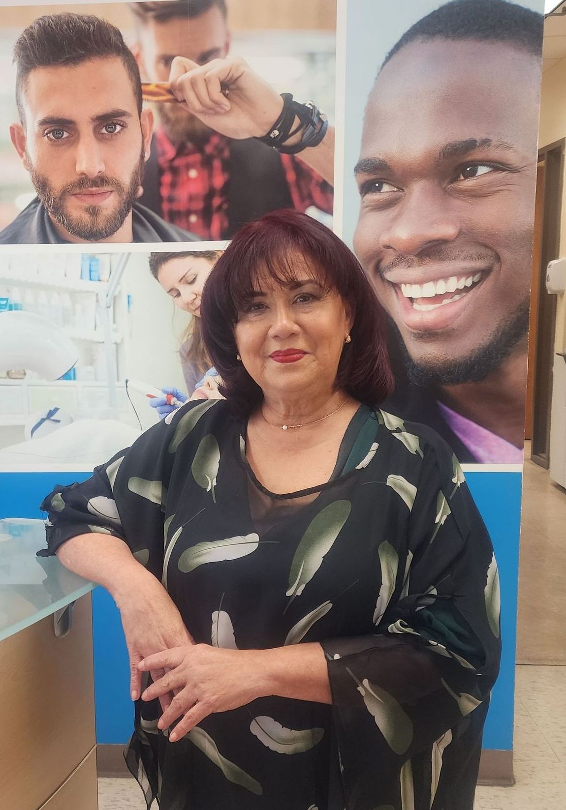 Woman with dark hair, smiling, in front of a clinic advertisement. She's wearing a black patterned top.
