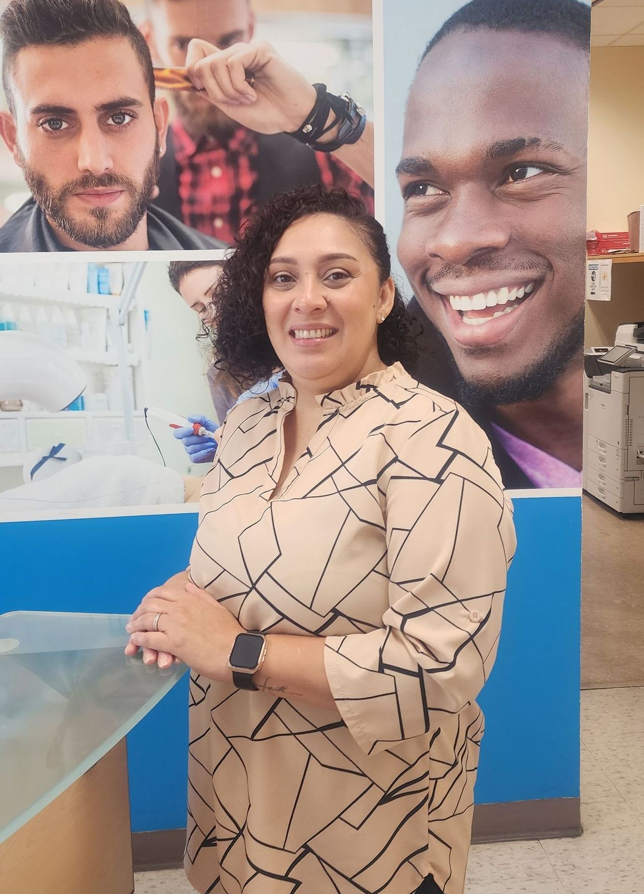 Woman in patterned shirt smiles, stands by a counter, in front of men's grooming posters.