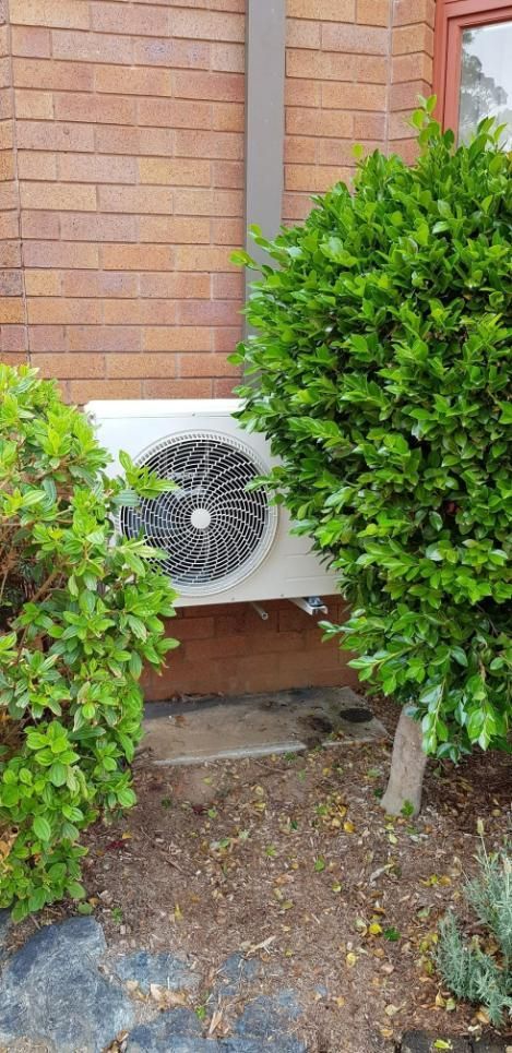 A Fan Is Sitting on The Side of A Brick Building Next to A Bush — Gleeson's Air Conditioning in Lake Cathie, NSW