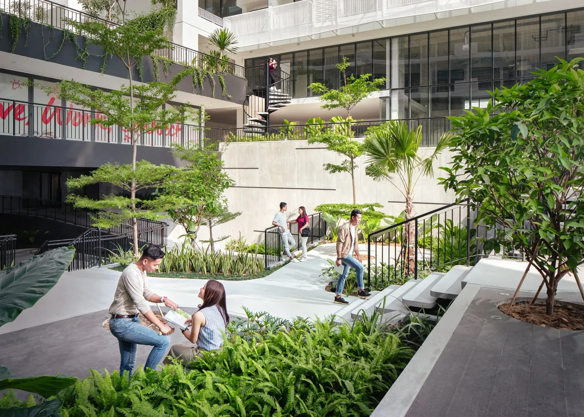 An outdoor courtyard with people walking, sitting, and surrounded by greenery. Buildings with large windows are in the background.