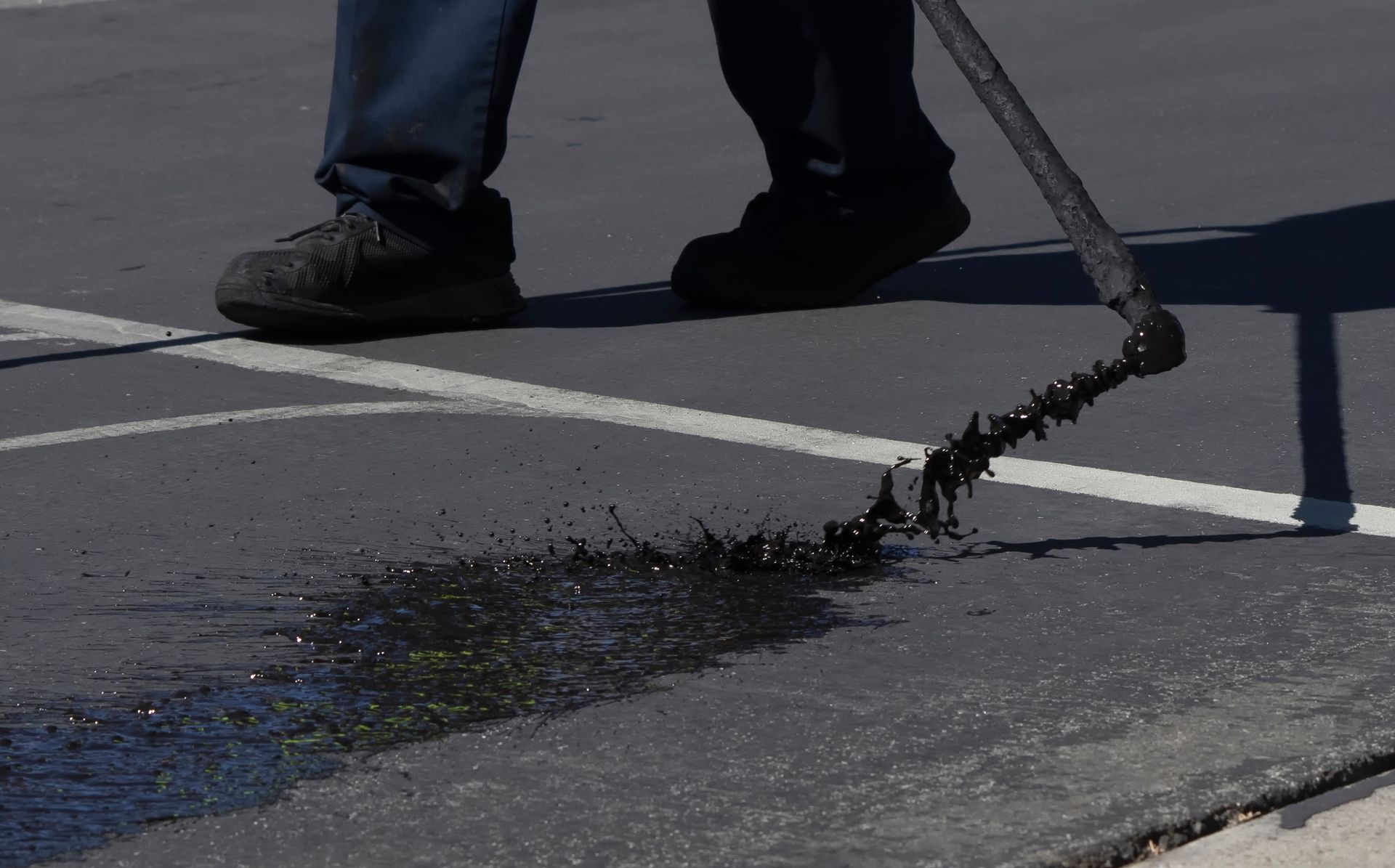 Worker scraping asphalt from a street with a long metal tool, leaving a pile of debris.