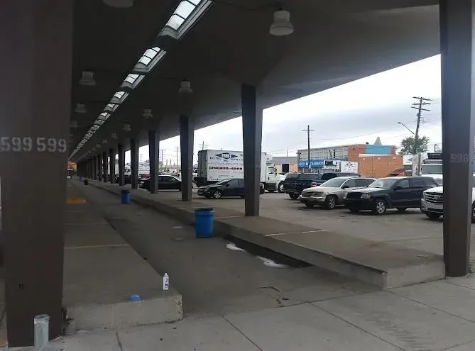 Covered outdoor parking area with vehicles and blue trash can. Gray and brown tones.